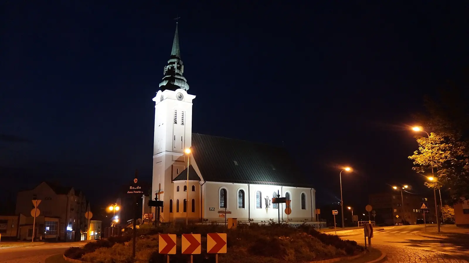 Oświetlony nocą biały kościół z wieżą przy skrzyżowaniu w centrum Miastka